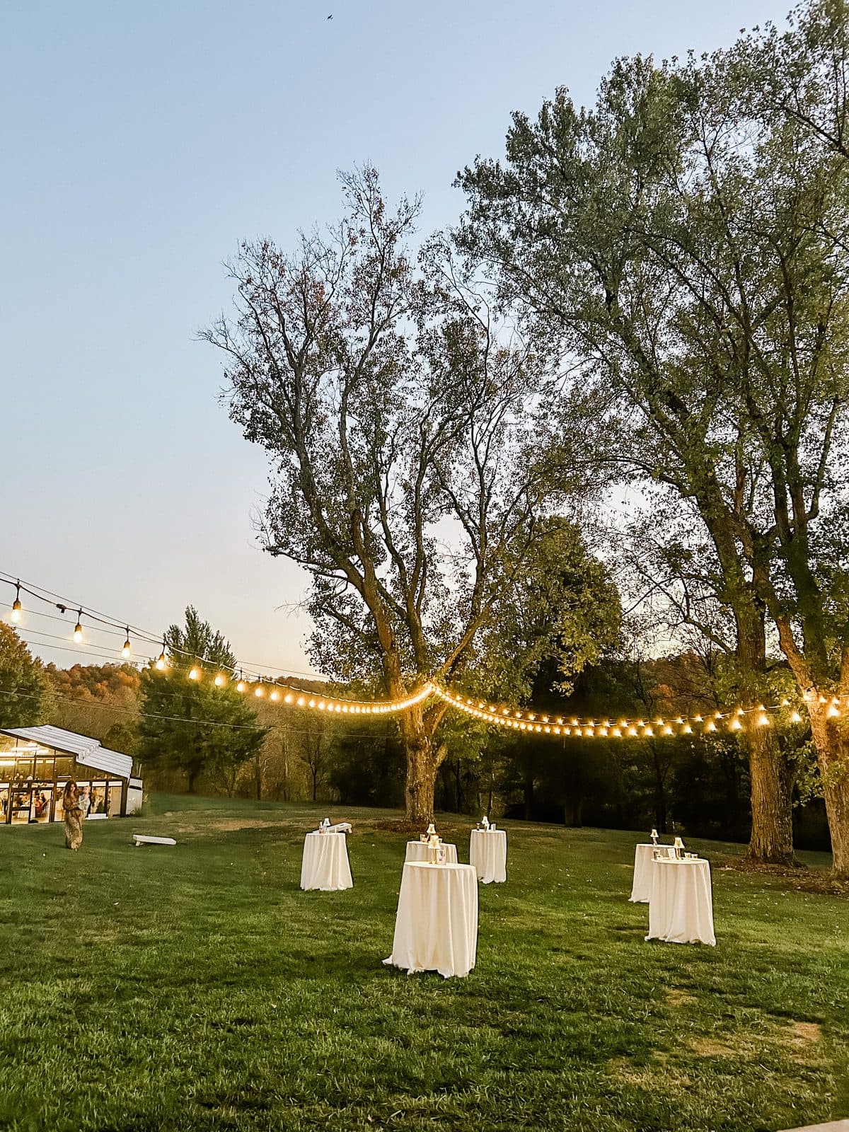 An outdoor lawn set with cocktail tables under string lights at dusk, a tent in the distance