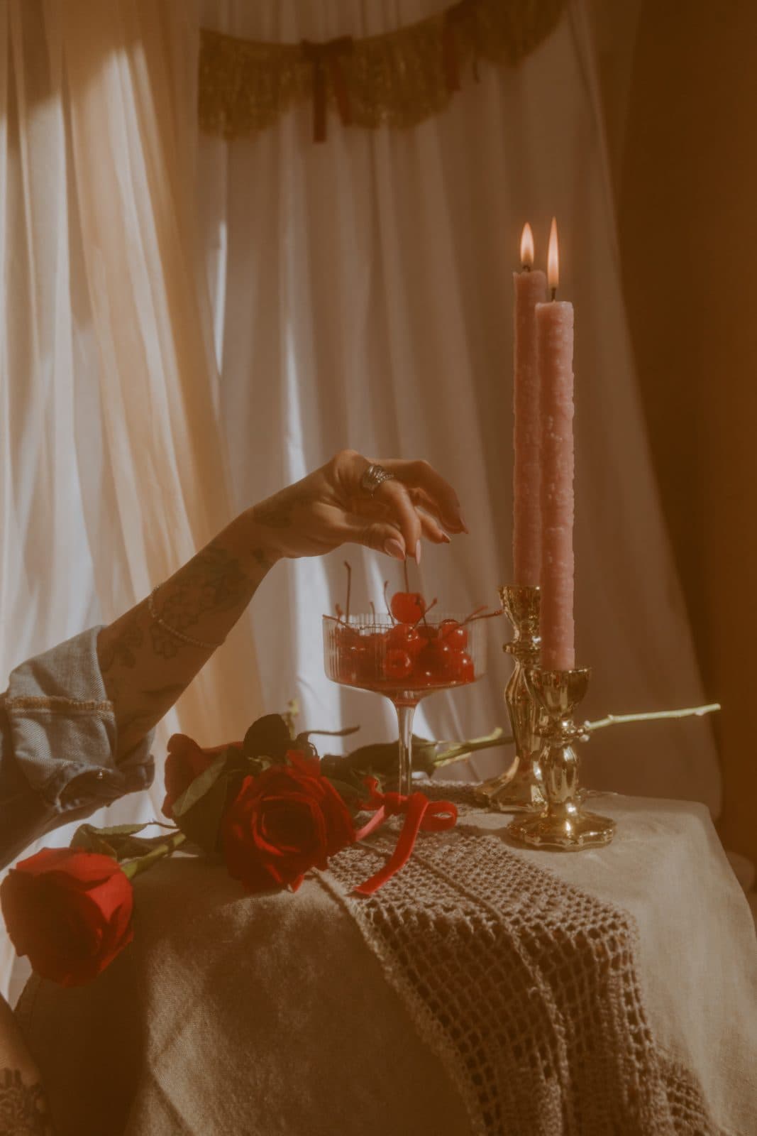 A hand picking a cherry from a coupe glass beside lit pink taper candles and red roses on a lace-draped table