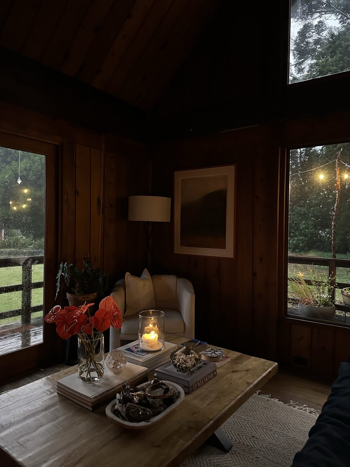 A cozy candlelit cabin sitting room with an armchair, a coffee table and string lights outside the windows