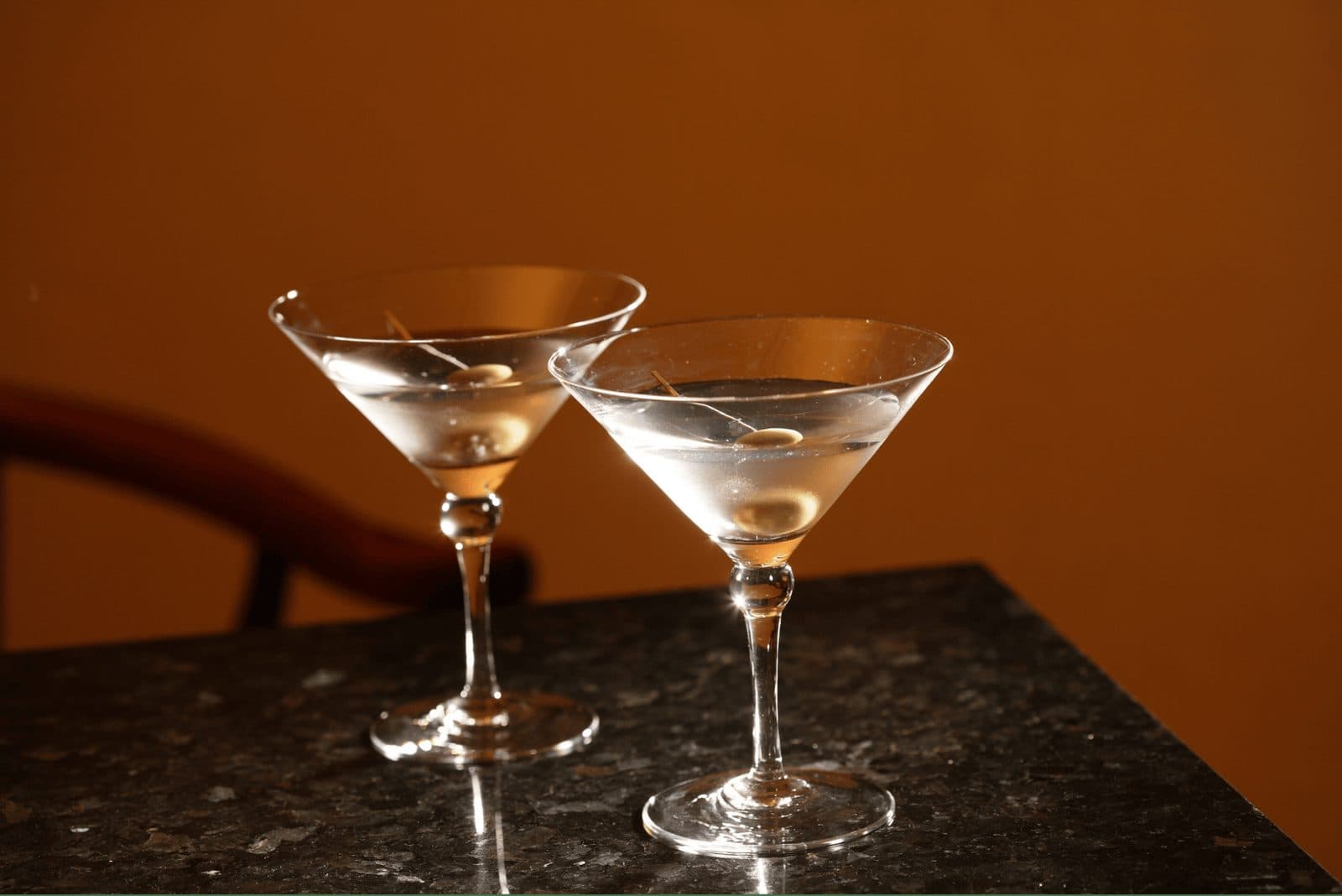 Two martini glasses side by side on a stone bar against an amber wall