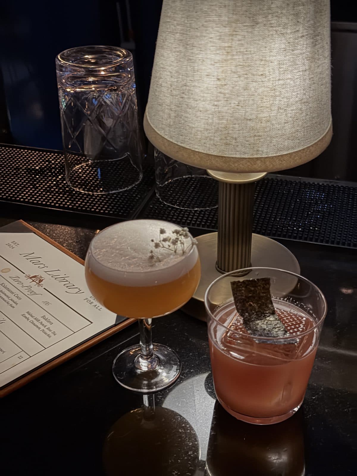A printed menu card beside two cocktails under a brass table lamp