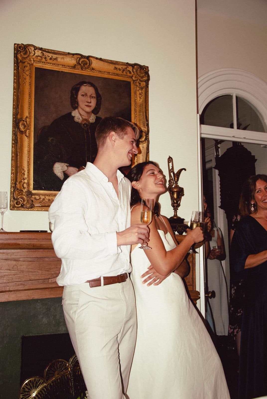 A couple in white embracing and laughing at their reception beside a gilt-framed portrait