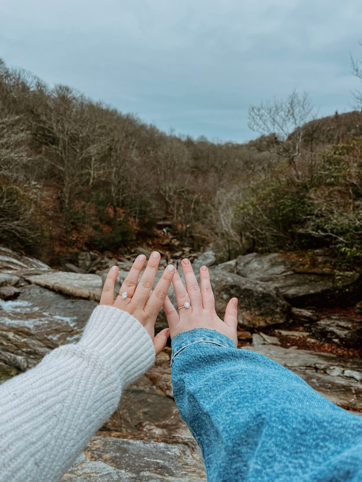 Two hands showing engagement rings in front of a rocky mountain stream and bare trees