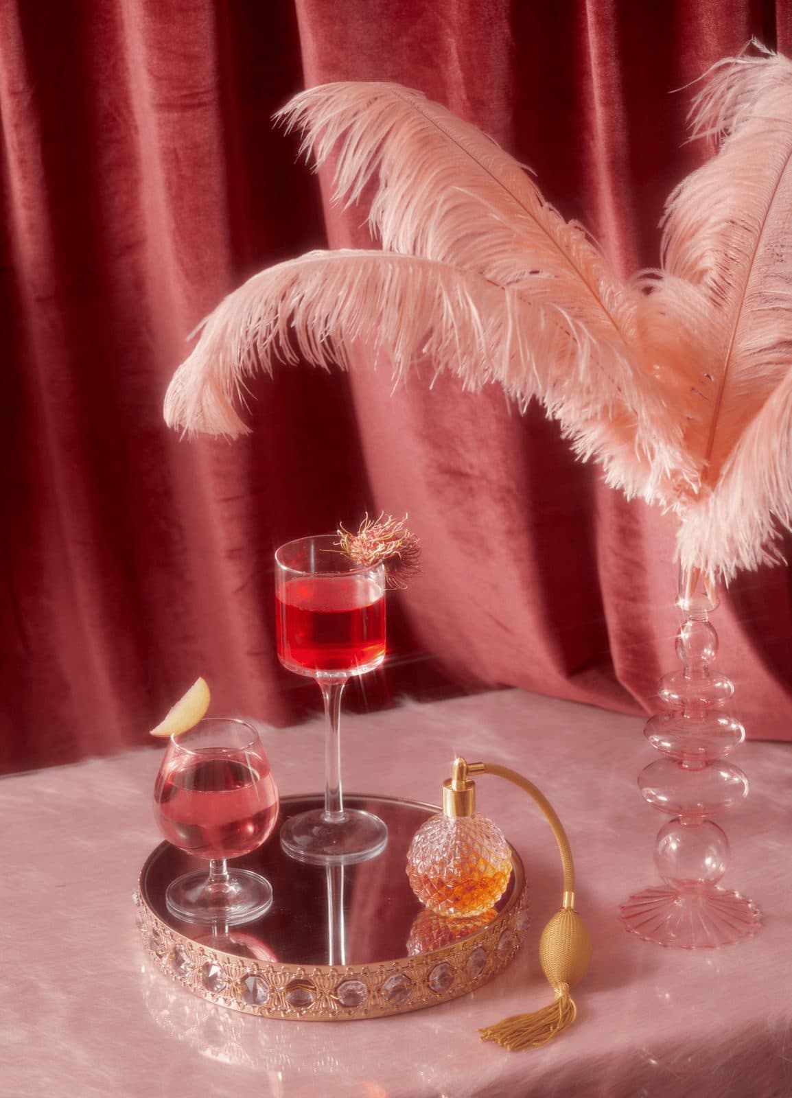 A styled vignette of pink ostrich feathers, a cocktail and a vintage perfume bottle on a mirrored tray