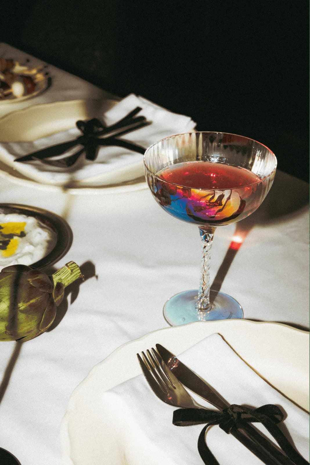 A place setting with black flatware tied with ribbon, a coupe glass and white linen